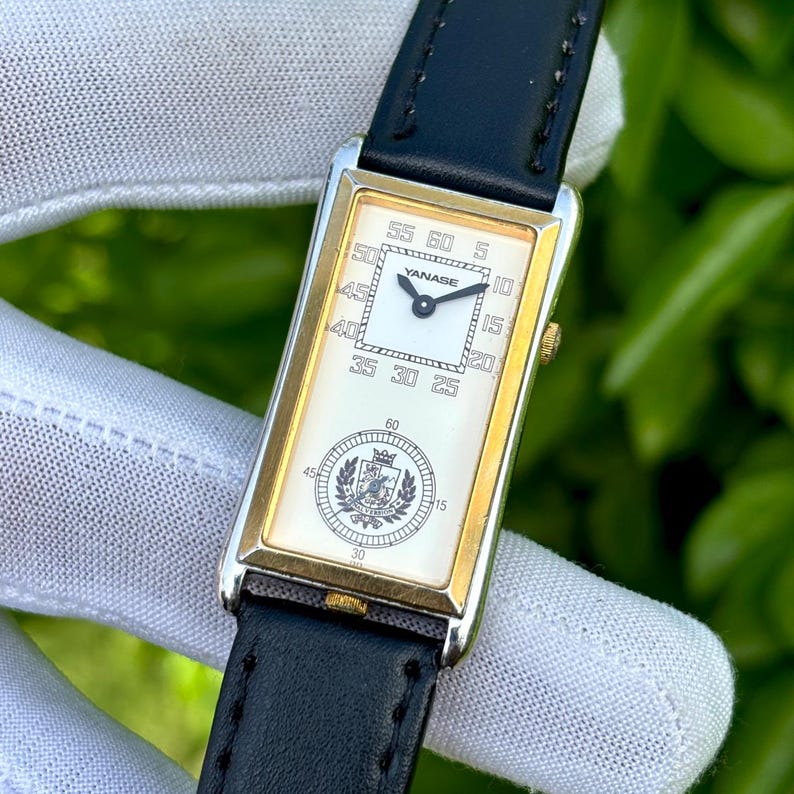 May include: Rectangular wristwatch with a gold-tone case and a white face. The watch features black hands, a black leather band, and the brand name "YANASE" on the face. The watch has a second dial with a crest.