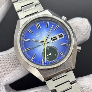 May include: A vintage Seiko 5 Sports Speed-Timer watch with a blue gradient dial. The watch features a stainless steel case and bracelet, a day-date display, and a subdial. The dial displays the text "SEIKO 5 SPORTS Speed-Timer".