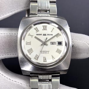 May include: A vintage Orient AAA Deluxe wristwatch with a silver-tone stainless steel band and case. The watch features a white face with Roman numeral hour markers, a date display, and the text "WATERPROOF 33 JEWELS".