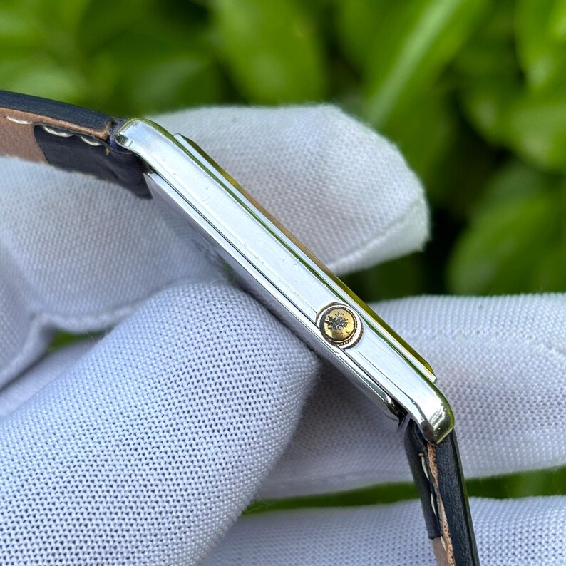 May include: A silver rectangular wristwatch with a black leather band. The watch has a gold-colored crown with a textured design. The watch is shown from a side angle, with the band partially visible.