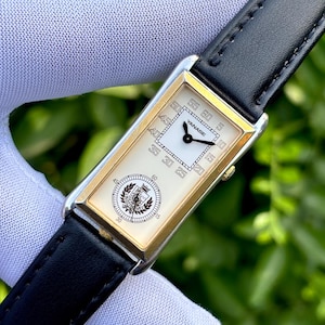 May include: A rectangular wristwatch with a white face and gold-colored trim. The watch has a black leather band and features the brand name "YANASE". The watch face displays the time with black hands and numerals.