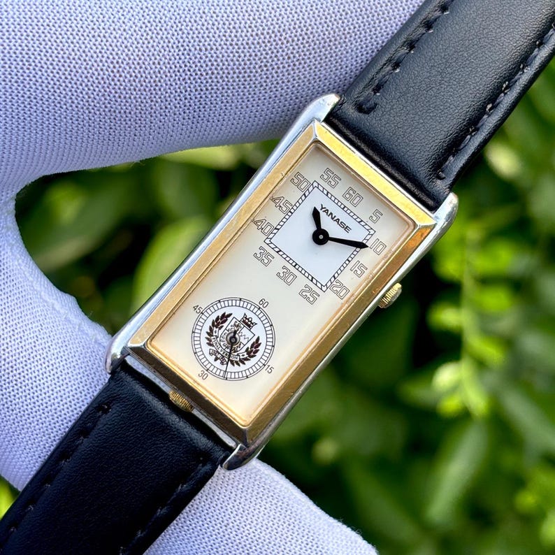 May include: A rectangular wristwatch with a white face and gold-colored trim. The watch has a black leather band and features the brand name "YANASE". The watch face displays the time with black hands and numerals.