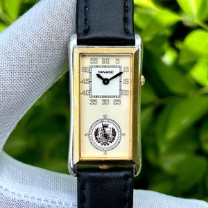 May include: Rectangular wristwatch with a cream-colored face, gold-tone bezel, and black leather band. The watch face features the brand name "YANASE" and a circular emblem. The watch displays the time with black hands and numerals.