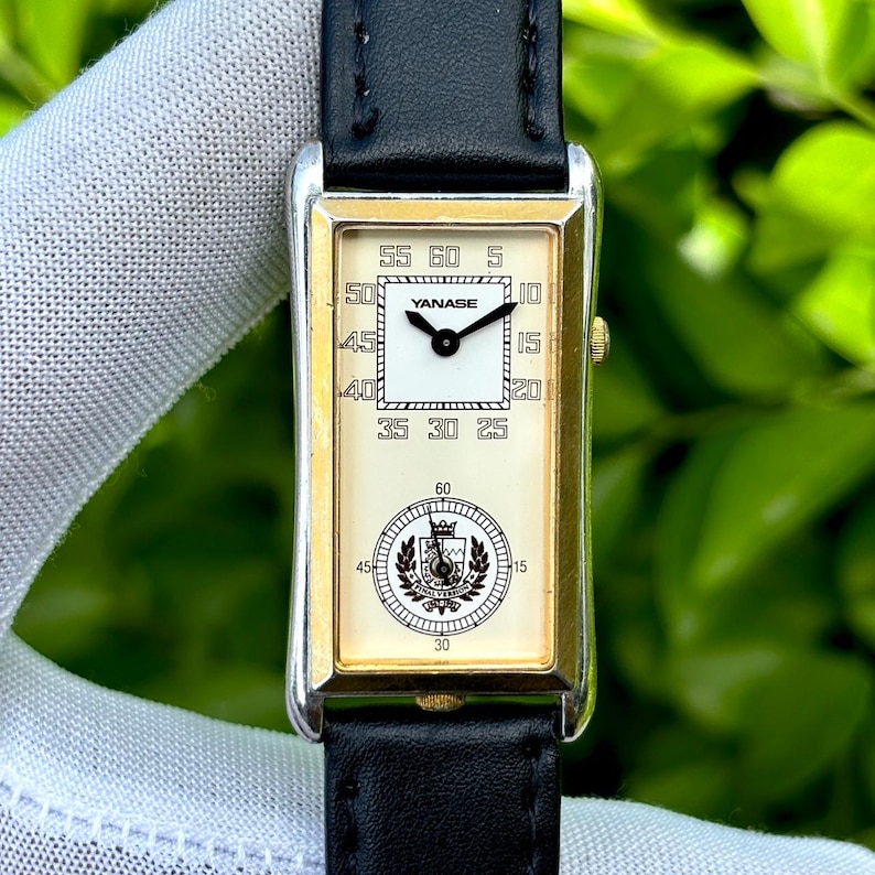 May include: Rectangular wristwatch with a cream-colored face, gold-tone bezel, and black leather band. The watch face features the brand name "YANASE" and a circular emblem. The watch displays the time with black hands and numerals.