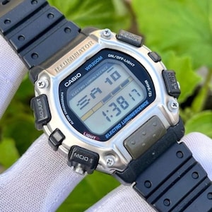 Può includere: Orologio digitale Casio con cinturino in resina nera e cassa color argento. Il quadrante mostra l'ora e la data, con il testo "ILLUMINATOR" e "ELECTRO LUMINESCENCE". L'orologio è impermeabile fino a 200 metri.