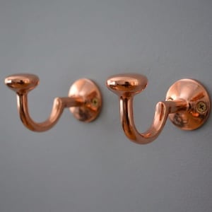 Puede incluir: Dos perchas de pared de color cobre con un diseño curvo. Cada gancho tiene una parte superior redondeada y un brazo curvo que se extiende hasta una base circular, montado en una pared gris. Los ganchos son de metal brillante.