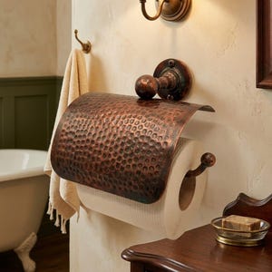 May include: A copper toilet roll holder with a hammered finish, featuring a matching knob and mounting plate. The holder is fixed to a light-toned wall in a bathroom. A roll of white toilet paper is in place.