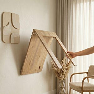 Puede incluir: Una mesa de madera plegable montada en la pared, con un acabado de madera clara, se está abriendo. La mesa está montada en una pared de color neutro, con una pieza de arte mural decorativa a la izquierda. Una mano está abriendo la mesa. La habitación tiene una combinación de colores neutros.