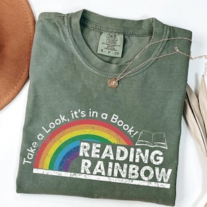 Op de afbeelding: Olijfgroen t-shirt met de tekst "Take a Look, it's in a Book! READING RAINBOW" over een regenboogafbeelding. Een gouden ketting met een hanger ligt op het shirt. Het shirt is op een wit oppervlak gevouwen.