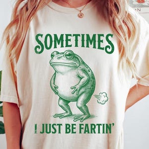 Peut inclure: Un t-shirt à manches courtes ivoire avec un motif vert représentant une grenouille debout. Le texte au-dessus de la grenouille indique "SOMETIMES" et le texte en dessous "I JUST BE FARTIN'" avec un petit nuage derrière la grenouille.