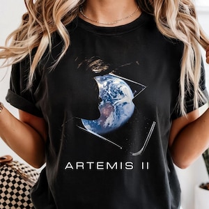 Könnte beinhalten: Schwarzes T-Shirt mit einer Grafik der Erde in einem geometrischen Design und dem Text "ARTEMIS II" in Weiß. Das Shirt hat kurze Ärmel und eine lockere Passform. Das Bild zeigt eine Person, die das Shirt trägt.