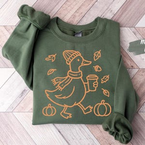 Pode incluir: Uma camisola de gola redonda verde militar com um gráfico de linhas cor de laranja de um pato com gorro e cachecol, segurando um copo de café, rodeado por folhas caídas e duas abóboras. O texto Military Green é visível no canto.