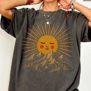 May include: A charcoal gray short-sleeve t-shirt featuring a graphic print of a yellow sun with a sleeping face, surrounded by radiating golden sunbeams above a mountain range with pine trees. The word Pepper is printed in the top right corner.