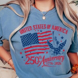K&ouml;nnte beinhalten: Hellblaues T-Shirt mit patriotischem Design. Die Grafik zeigt eine rot-blaue amerikanische Flagge, die Freiheitsstatue und einen Adler. Der Text lautet "UNITED STATES OF AMERICA 250th Anniversary 1776-2026."