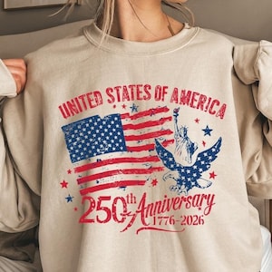 K&ouml;nnte beinhalten: Beiges Sweatshirt mit patriotischem Design. Auf der Vorderseite steht der Text "UNITED STATES OF AMERICA" &uuml;ber einer stilisierten amerikanischen Flagge, einem Adler und der Freiheitsstatue. Darunter steht "250th Anniversary 1776-2026".