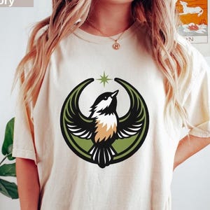 Può includere: T-shirt color avorio con un disegno di uccello. La grafica include un uccello con le ali spiegate, una stella e una cornice circolare verde e nera. La maglietta ha una vestibilità ampia.