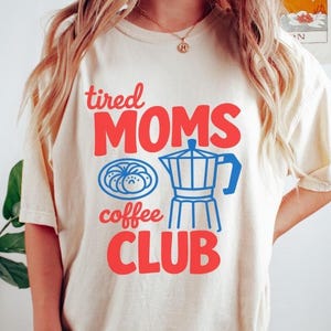 Könnte beinhalten: Elfenbeinfarbenes T-Shirt mit dem Text "tired MOMS coffee CLUB" in Rot und Blau. Das Design beinhaltet eine blaue Kaffeemaschine und ein blaues Gebäck. Das Shirt ist ein kurzärmeliger, lässiger Stil.