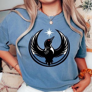 May include: Bluejean-coloured t-shirt featuring a black and white graphic of a bird with outstretched wings, a star, and a circular design. The bird has a red eye. The shirt is a solid colour and has short sleeves.