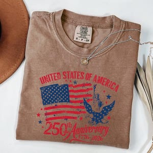 K&ouml;nnte beinhalten: Braunes T-Shirt mit einer Grafik der amerikanischen Flagge, der Freiheitsstatue und einem Adler. Der Text lautet "UNITED STATES OF AMERICA" und "250th Anniversary 1776-2026". Eine silberne Halskette mit Anh&auml;nger liegt auf dem Shirt.