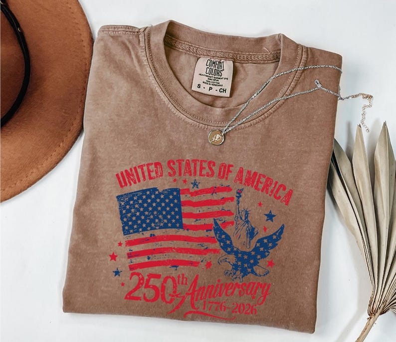K&ouml;nnte beinhalten: Braunes T-Shirt mit einer Grafik der amerikanischen Flagge, der Freiheitsstatue und einem Adler. Der Text lautet "UNITED STATES OF AMERICA" und "250th Anniversary 1776-2026". Eine silberne Halskette mit Anh&auml;nger liegt auf dem Shirt.