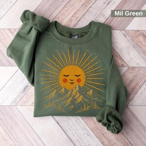 May include: A military green crewneck sweatshirt featuring a graphic print of a golden sun with a sleeping face and rosy cheeks rising above a mountain range with pine trees. The text Mil Green is printed in the upper right corner of the image.