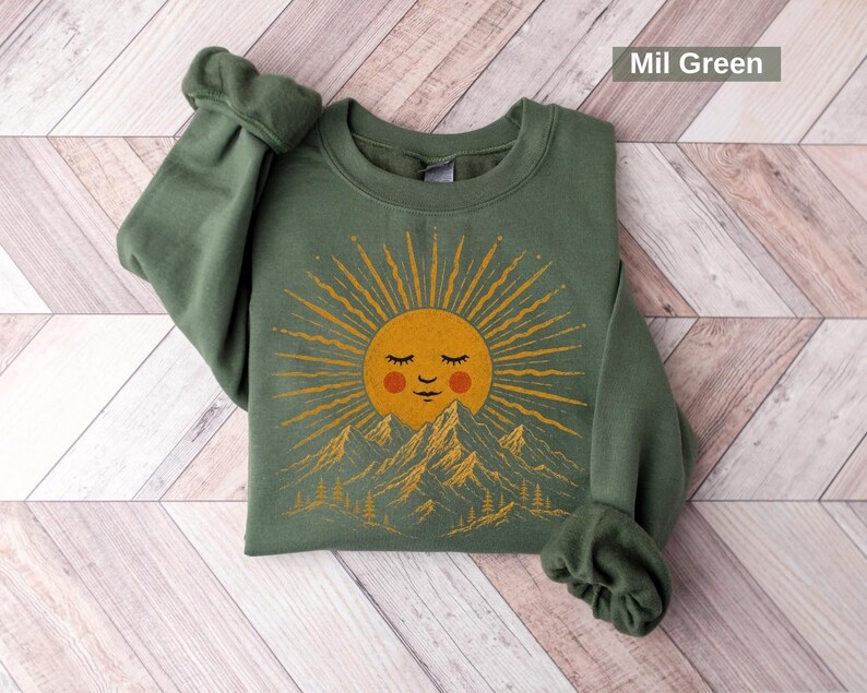May include: A military green crewneck sweatshirt featuring a graphic print of a golden sun with a sleeping face and rosy cheeks rising above a mountain range with pine trees. The text Mil Green is printed in the upper right corner of the image.