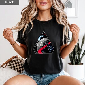 Op de afbeelding: Een zwart t-shirt met korte mouwen en een driehoekige afbeelding op de borst. Het ontwerp bevat een maan, een gedeeltelijk zicht op de aarde, een rode boog en de letter A, met de tekst WISEMAN GLOVER KOCH HANSEN onderaan gedrukt.