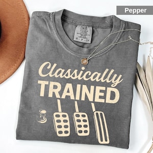 May include: A pepper-colored, garment-dyed t-shirt featuring the text Classically TRAINED in cream-colored lettering. Below the text is a graphic illustration of a clutch, brake, and gas pedal set. The shirt has a crew neckline and a tag.