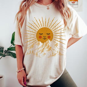 May include: An ivory short-sleeve t-shirt featuring a graphic print of a golden sun with a smiling face, positioned above a mountain range with pine trees. The sun has radiating rays and red circular cheeks. The shirt has a crew neckline.