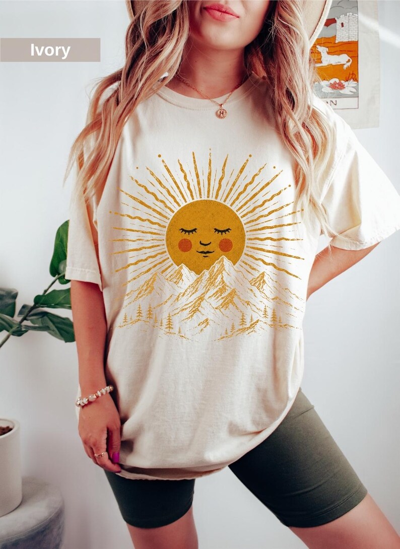 May include: An ivory short-sleeve t-shirt featuring a graphic print of a golden sun with a smiling face, positioned above a mountain range with pine trees. The sun has radiating rays and red circular cheeks. The shirt has a crew neckline.