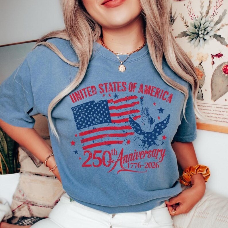 K&ouml;nnte beinhalten: Hellblaues T-Shirt mit patriotischem Design. Die Grafik zeigt eine rot-blaue amerikanische Flagge, die Freiheitsstatue und einen Adler. Der Text lautet "UNITED STATES OF AMERICA 250th Anniversary 1776-2026."
