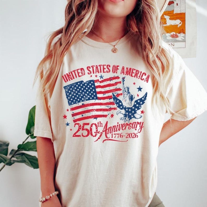 K&ouml;nnte beinhalten: Cremefarbenes T-Shirt mit patriotischem Design. Die Grafik zeigt die amerikanische Flagge, einen Adler und die Freiheitsstatue mit dem Text "United States of America" und "250th Anniversary 1776-2026".