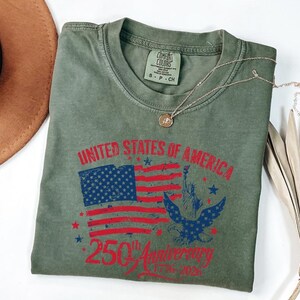 K&ouml;nnte beinhalten: Olivgr&uuml;nes T-Shirt mit einer Grafik der amerikanischen Flagge, der Freiheitsstatue und einem Adler. Der Text lautet "UNITED STATES OF AMERICA 250th Anniversary 1776-2026". Eine goldene Halskette liegt auf dem T-Shirt.