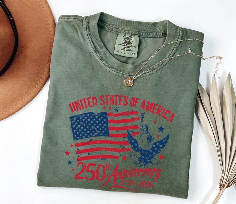 K&ouml;nnte beinhalten: Olivgr&uuml;nes T-Shirt mit einer Grafik der amerikanischen Flagge, der Freiheitsstatue und einem Adler. Der Text lautet "UNITED STATES OF AMERICA 250th Anniversary 1776-2026". Eine goldene Halskette liegt auf dem T-Shirt.