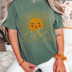 May include: A moss green short-sleeve t-shirt featuring a graphic print of a smiling sun with closed eyes and rosy cheeks positioned above a mountain range and pine trees. The sun and landscape design is rendered in a golden yellow color.