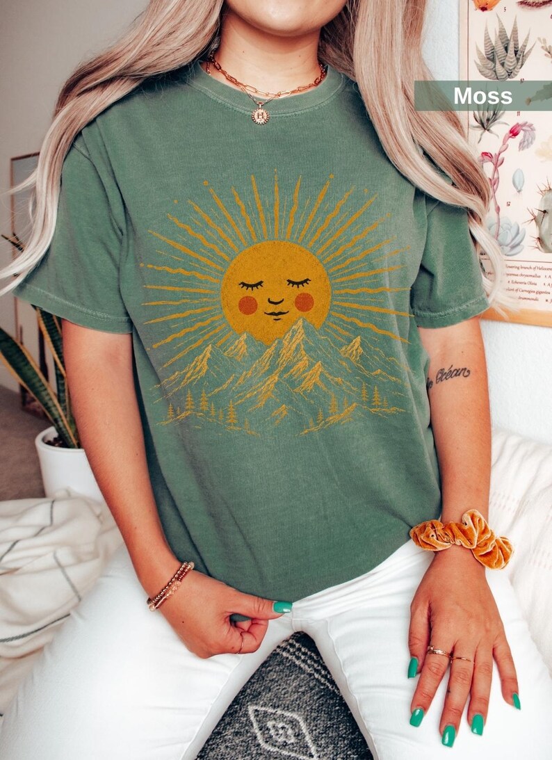 May include: A moss green short-sleeve t-shirt featuring a graphic print of a smiling sun with closed eyes and rosy cheeks positioned above a mountain range and pine trees. The sun and landscape design is rendered in a golden yellow color.