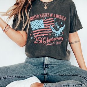 K&ouml;nnte beinhalten: Dunkelgraues T-Shirt mit einem verblassten Design der amerikanischen Flagge, dem Text "UNITED STATES OF AMERICA" und "250th Anniversary 1776-2026". Das Shirt zeigt auch die Freiheitsstatue und einen Adler.