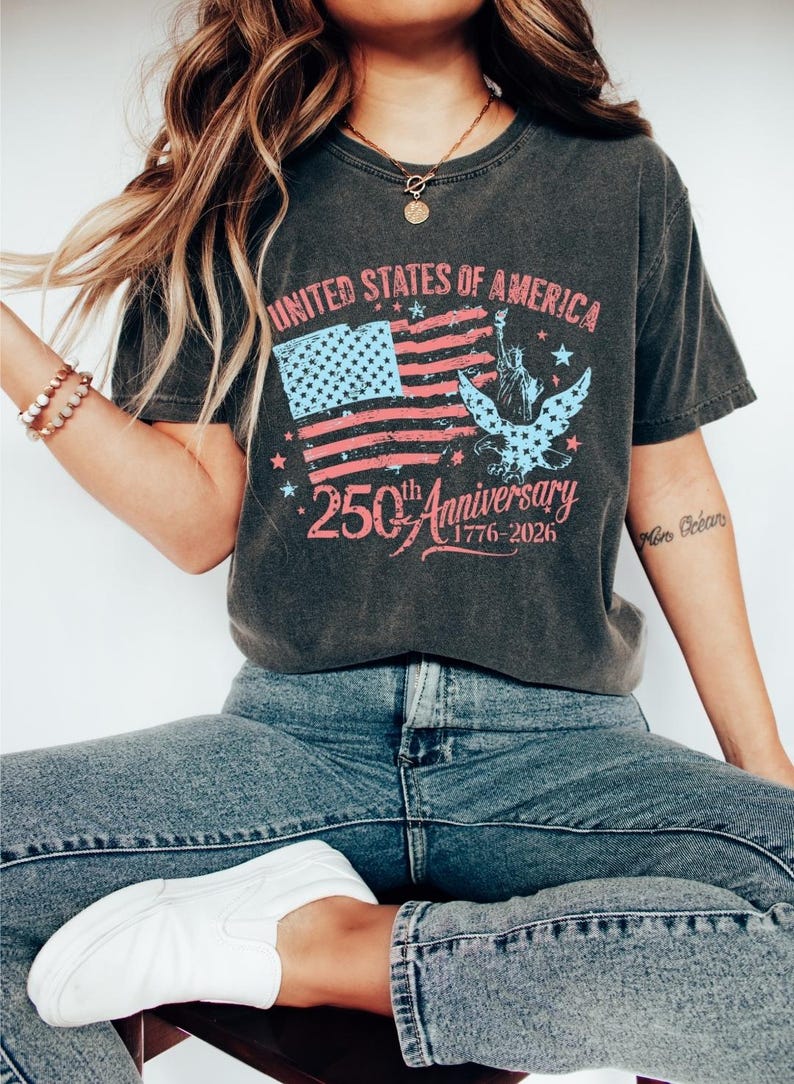 K&ouml;nnte beinhalten: Dunkelgraues T-Shirt mit einem verblassten Design der amerikanischen Flagge, dem Text "UNITED STATES OF AMERICA" und "250th Anniversary 1776-2026". Das Shirt zeigt auch die Freiheitsstatue und einen Adler.