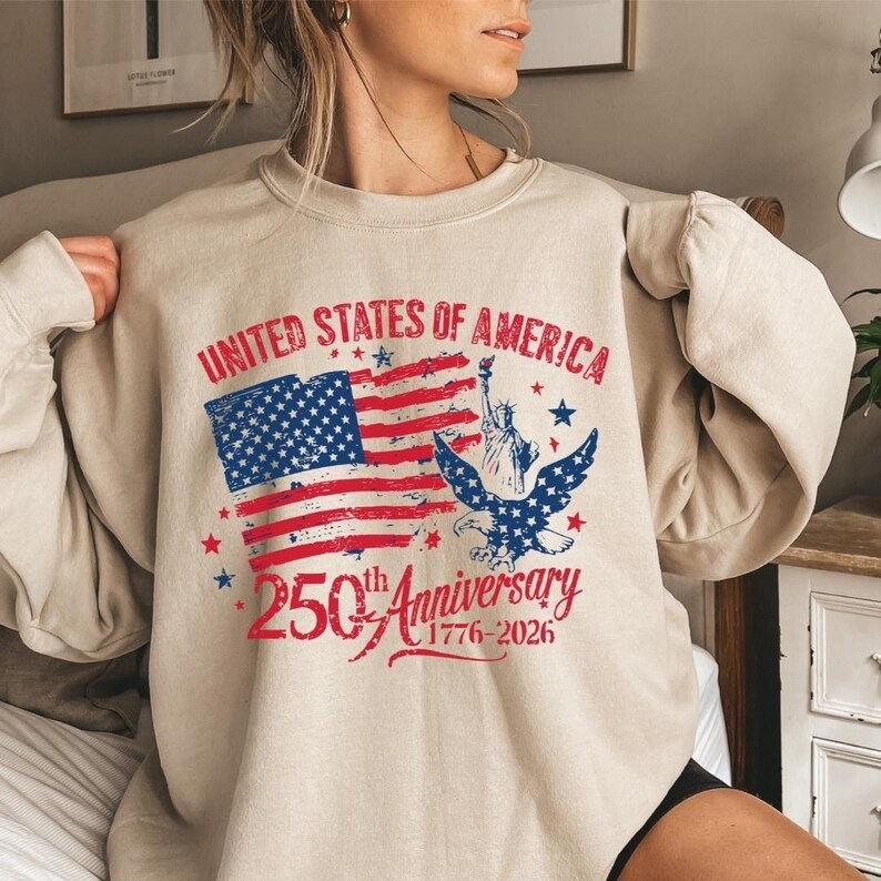 K&ouml;nnte beinhalten: Beiges Sweatshirt mit patriotischem Design. Auf der Vorderseite steht der Text "UNITED STATES OF AMERICA" &uuml;ber einer stilisierten amerikanischen Flagge, einem Adler und der Freiheitsstatue. Darunter steht "250th Anniversary 1776-2026".