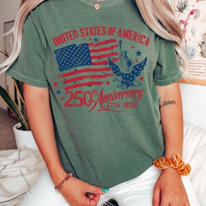 K&ouml;nnte beinhalten: Olivgr&uuml;nes T-Shirt mit einem Grafikdesign zum 250. Jahrestag der Vereinigten Staaten von Amerika. Das Design zeigt die amerikanische Flagge, die Freiheitsstatue und einen Adler. Der Text lautet: "UNITED STATES OF AMERICA 250th Anniversary 1776-2026."