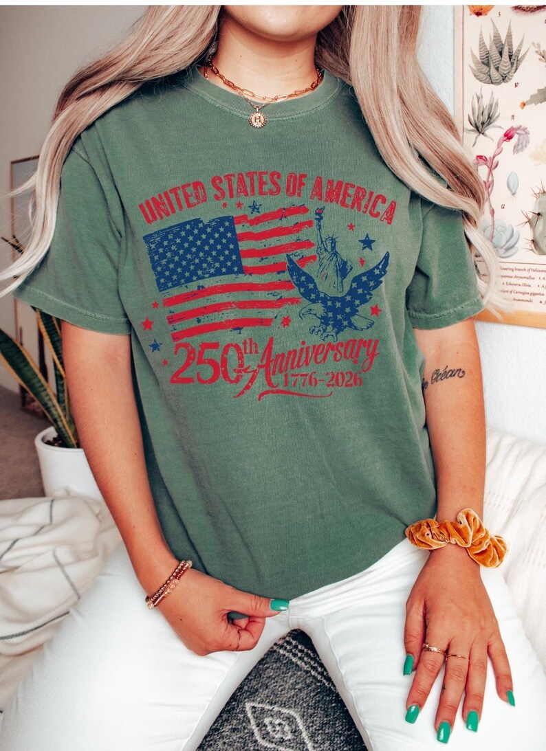 K&ouml;nnte beinhalten: Olivgr&uuml;nes T-Shirt mit einem Grafikdesign zum 250. Jahrestag der Vereinigten Staaten von Amerika. Das Design zeigt die amerikanische Flagge, die Freiheitsstatue und einen Adler. Der Text lautet: "UNITED STATES OF AMERICA 250th Anniversary 1776-2026."
