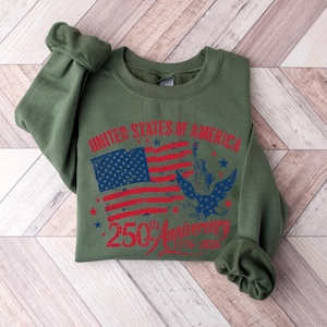K&ouml;nnte beinhalten: Ein olivgr&uuml;nes Sweatshirt mit einem Grafikdesign zum 250. Jahrestag der Vereinigten Staaten von Amerika. Das Design umfasst eine verblasste amerikanische Flagge, die Freiheitsstatue und einen Adler.