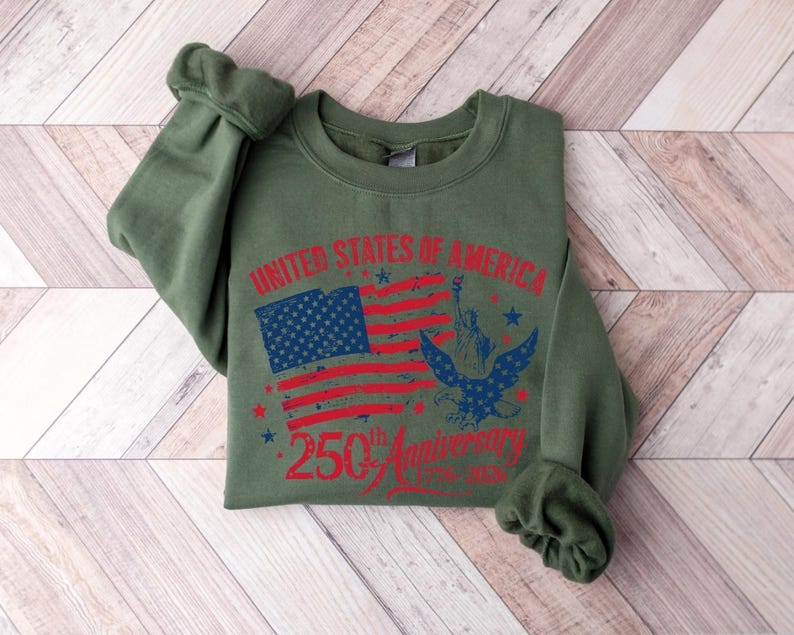 K&ouml;nnte beinhalten: Ein olivgr&uuml;nes Sweatshirt mit einem Grafikdesign zum 250. Jahrestag der Vereinigten Staaten von Amerika. Das Design umfasst eine verblasste amerikanische Flagge, die Freiheitsstatue und einen Adler.