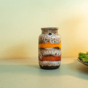 Op de afbeelding: Een vintage keramische vaas met een cilindrische vorm. De vaas heeft horizontale banden in oranje, wit en bruin. De vaas is ongeveer 15 cm hoog en heeft een getextureerd oppervlak. Een decoratief item voor woondecoratie.