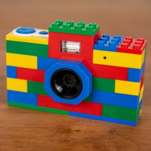 Könnte beinhalten: Eine bunte Kamera aus ineinandergreifenden Spielzeugbausteinen. Die Kamera hat ein schwarzes Objektiv, einen Blitz und Knöpfe oben. Der Körper besteht aus roten, gelben, grünen und blauen Steinen, was ein verspieltes Design ergibt. Die Kamera steht auf einer Holzoberfläche.