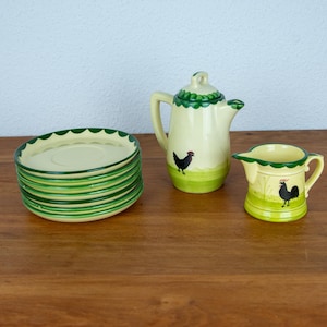 Pode incluir: Um conjunto de pratos creme com bordas verdes onduladas, um creme correspondente e um jarro pequeno. Cada peça apresenta um desenho de galo preto sobre uma base verde clara. A chaleira tem uma tampa verde.
