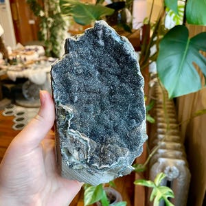 Puede incluir: Una geoda grande, de color gris oscuro, con una superficie texturizada y cristalina. La geoda tiene áreas de color verde oscuro y blanco, con una forma natural rugosa. El objeto se sostiene en una mano, mostrando su tamaño y detalle. Un artículo de decoración natural.