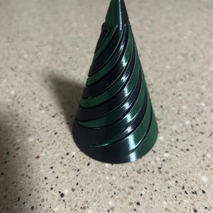 Puede incluir: Objeto cónico impreso en 3D con un diseño en espiral. El objeto es principalmente verde oscuro con bandas negras alternas. El cono se estrecha hasta un punto en la parte superior. El objeto está sobre una superficie clara y moteada.