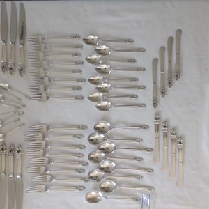 May include: An assortment of silver-coloured cutlery, including knives, forks, and spoons, laid out on a white surface. The silverware features decorative handle detailing. The arrangement implies a formal dining setting, with several place settings visible.