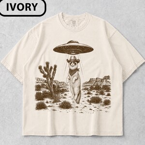 Puede incluir: Una camiseta color marfil con un estampado gráfico marrón de un gato con sombrero de vaquero y pañuelo, abducido por un OVNI en un paisaje desértico con un árbol de Josué. La palabra IVORY aparece en un recuadro negro en la esquina superior.